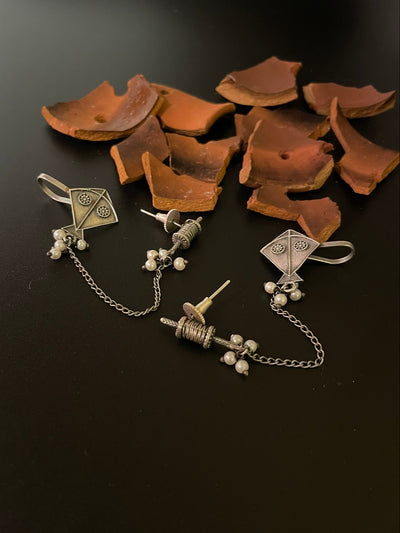 This image displays a pair of Kite Climber Stud Earrings, elegantly crafted with an oxidized silver finish. The earrings are designed in the shape of kites, featuring intricate details and a chain of pearls that add sophistication to the design.